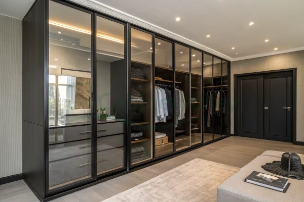 Luxury black wardrobe with soft-glow lighting and framed tinted glass doors, arranged along a textured wall in a spacious bedroom with a seating area and dual-access storage.